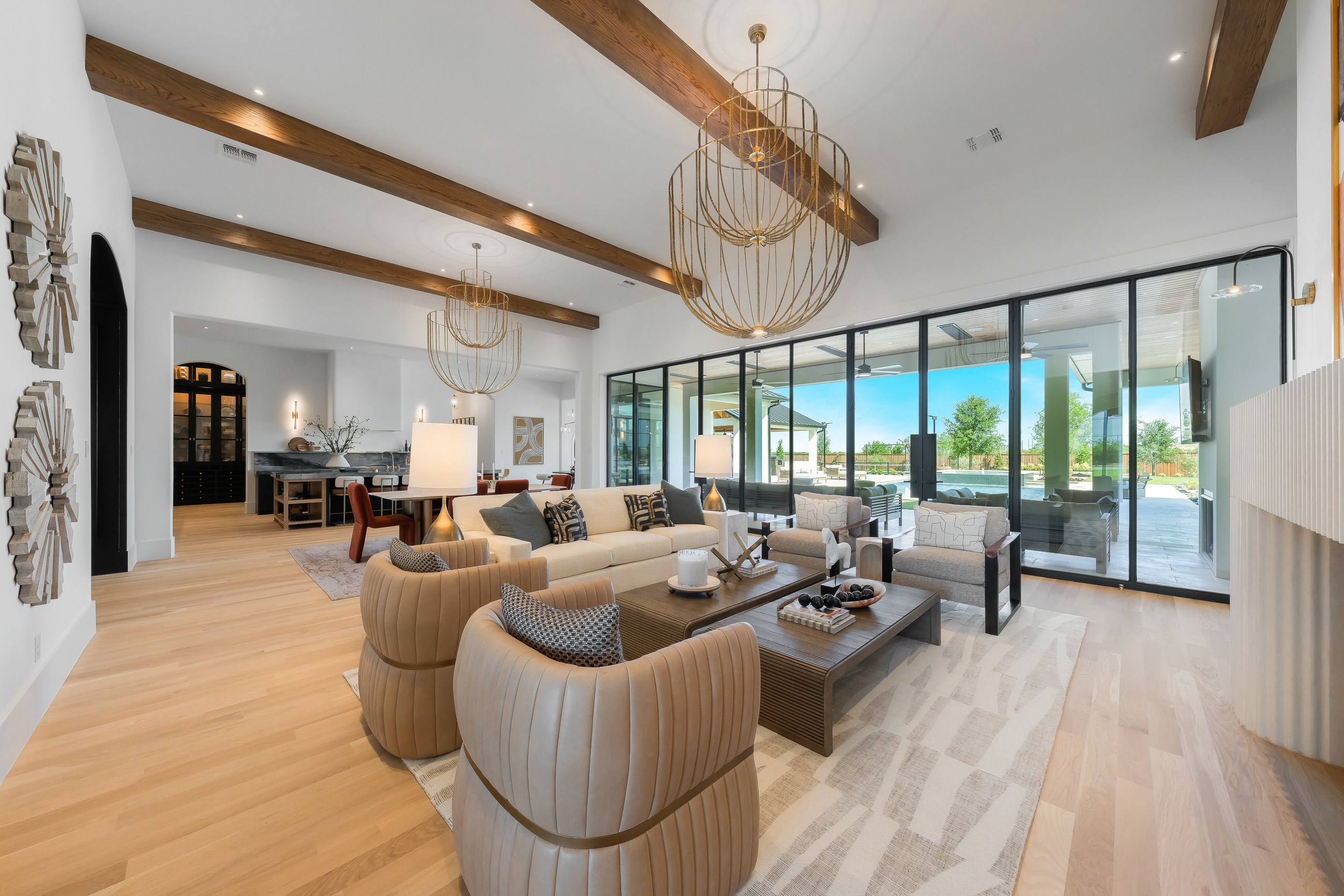 Great room with exposed wood beams, a brass chandelier and a full glass wall overlooking the grounds